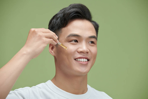 close up of young Asian man applying moisturizing serum isolated on green - Foto, afbeelding