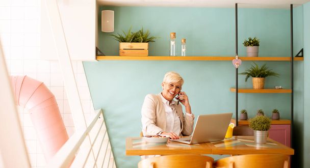 Starsza kobieta korzystająca z telefonu komórkowego podczas pracy na laptopie i picia świeżego soku pomarańczowego w kawiarni - Zdjęcie, obraz