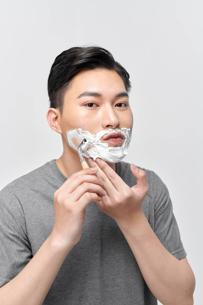 Positive attractive young guy with gel foam on lower face shaving with razor at mirror - Fotoğraf, Görsel