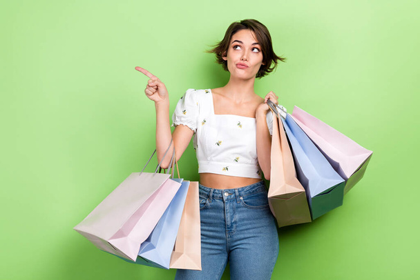 Photo of shiny unsure lady wear white crop top pointing finger looking empty space rising shoppers isolated green color background. - Фото, изображение