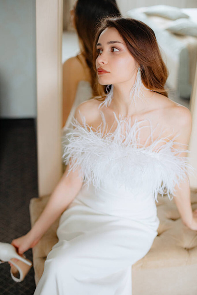 Charming young bride sitting on beige sofa next to mirror in wedding dress without shoulders adorned with feathers and holding high heels shoe in hand. Lady with brown hair wearing tassel earrings. - Foto, Imagem