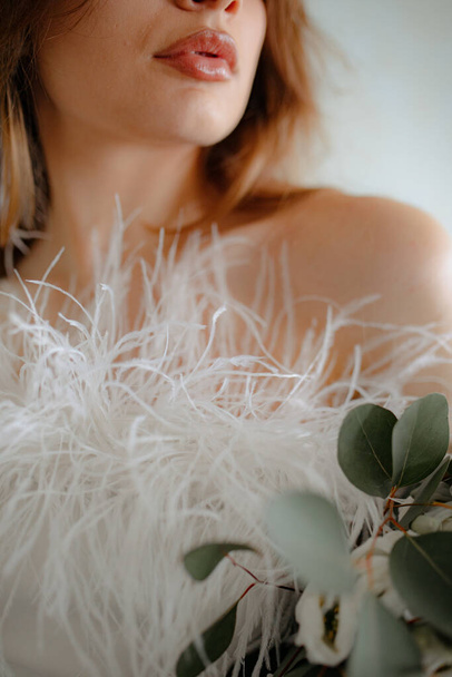 Close up of dark-haired young woman who dressed in shoulderless white clothes adorned with fluffy and little feathers. Bride with reddish brown lips holding flowers in hands and looking away. - 写真・画像