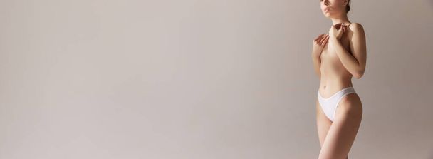 Image recadrée de jeune fille mince couvrant le sein avec la main. Modèle posant en sous-vêtements sur fond de studio gris. Mammoplastie. Concept de beauté naturelle, soins du corps et de la peau, santé, bien-être. Publicité - Photo, image