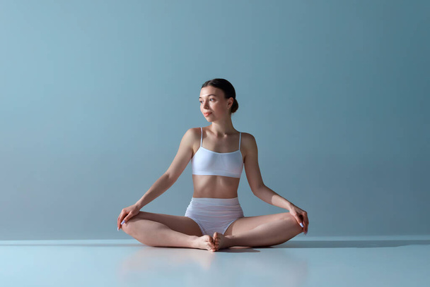 Portrait of beautiful, relaxed, young girl with slim body sitting in lotus pose in underwear on blue studio background. Concept of natural beauty, body and skin care, health, wellness, femininity. Ad - Photo, Image