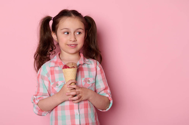 Белая очаровательная маленькая девочка держит мороженое в вафельном конусе, смотрит в сторону рекламного места на изолированном розовом фоне. Детей. Счастливого детства. Еда и напитки. Летняя концепция - Фото, изображение