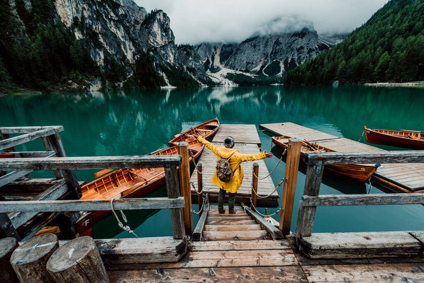 Rear view portrait of a hiker with arms outstretched visiting alpine lake at Braies, Italy. Happy hiker wearing yellow jacket and backpack enjoy the nature landscape at autumn. Wanderlust and travel. - Photo, Image