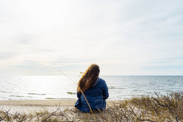 Femme aux cheveux longs et aux cheveux bouclés en veste bleue assise sur le rivage de la mer Baltique. - Photo, image