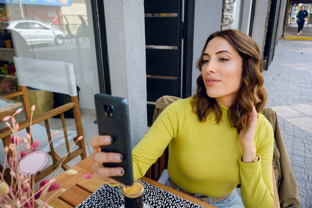 front view of young beautiful latina woman of argentinian ethnicity, taking a photo with her phone while sitting outside the restaurant, copy space. - Fotó, kép