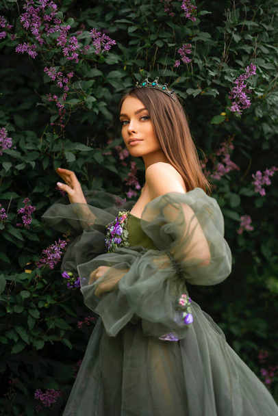 Linda rainha em coroa e belo vestido longo verde com flores posando no jardim lilás. Retratos de moda modelo jovem. - Foto, Imagem