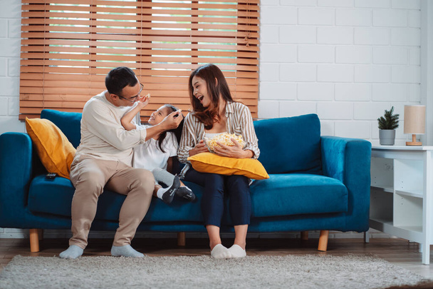 両親、幸せな娘、テレビを見ているか、自宅のリビングルームでソファの上でポップコーンを食べる映画で構成されるアジアの家族。リラックスした幸せ. - 写真・画像