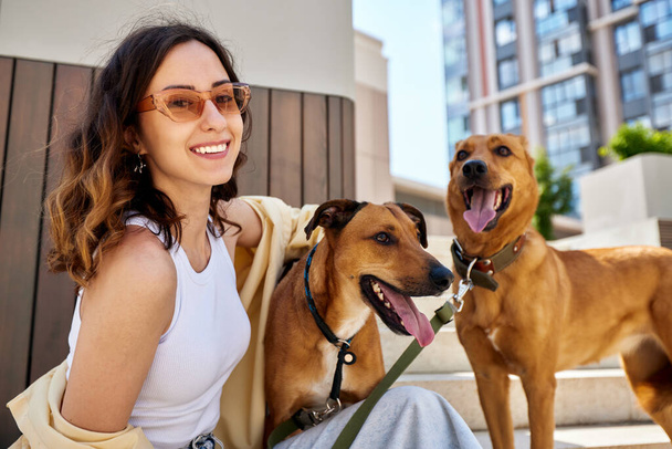 Charmante junge lächelnde Mädchen auf einem Spaziergang mit zwei goldfarbenen Hunden im Freien an einem sonnigen Tag. Das Mädchen umarmt Haustiere. Liebe zwischen Besitzer und Haustier. Aufzucht von Haustieren aus einem Tierheim. Nahaufnahme - Foto, Bild