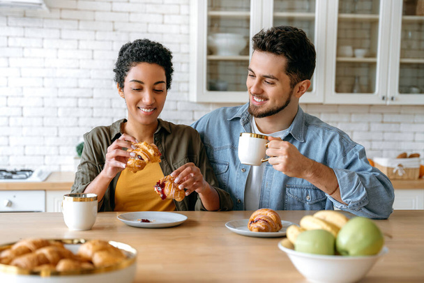 Huomenta aamiainen. Onnellinen monirotuinen pari, valkoihoinen mies ja afrikkalainen amerikkalainen nainen, viettää aikaa yhdessä kotona, he juovat kahvia, syövät croissanteja, keskustelevat eri aiheista, suunnittelevat vapaa-aikaa, hymyilevät - Valokuva, kuva