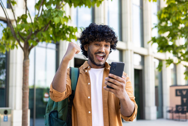 Young hispanic student celebrating victory and happy good news online, man reading page interent received notification and message, hold phone, use app win and good achievement. - Foto, imagen