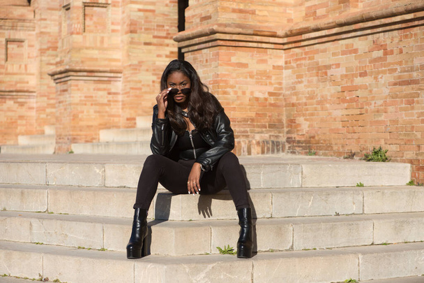 giovane e bella donna latina nera che indossa vestiti neri e occhiali da sole è seduto sui gradini della piazza più importante della città di seville, in Spagna. La foto è scattata dalla parte anteriore. - Foto, immagini
