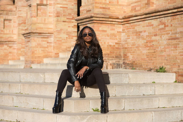 giovane e bella donna latina nera che indossa vestiti neri e occhiali da sole è seduto sui gradini della piazza più importante della città di seville, in Spagna. La foto è scattata dalla parte anteriore. - Foto, immagini