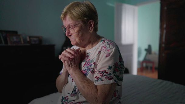 Religieuze Oudere vrouw zit naast haar bed in het gebed. HOPEFUL oudere dame biddend tot GOD in slaapkamer, authentieke echte huiselijke levensstijl beeltenis van ouderdom in GELOOF - Foto, afbeelding