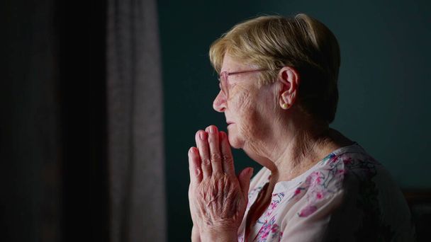 Ältere Frau, die am Fenster steht und zu Gott betet und Kreuzzeichen macht, katholische ältere Person in tiefer Andacht. Konzept Hoffnung und Glaube - Foto, Bild