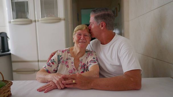 Yetişkin oğlunun, mutfakta otururken yaşlı anneyi alnının ortasından öpmesi arasında sevgi dolu bir aile anı. Otantik sevecen gerçek insanlar sahnesi - Fotoğraf, Görsel