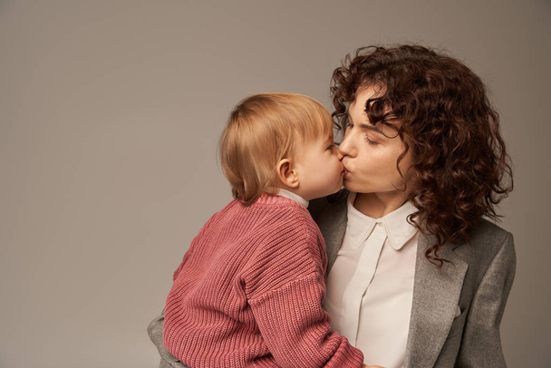 work life balance concept, curly woman in suit kissing her daughter on grey background, loving mother, balanced lifestyle, modern working woman. modern parenting, career and family  - 写真・画像