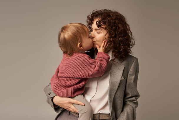 koncepcja równowagi życia zawodowego, kręcona kobieta w garniturze całująca córkę na szarym tle, kochająca matka, zrównoważony styl życia, nowoczesna kobieta pracująca. nowoczesne rodzicielstwo, więź i miłość - Zdjęcie, obraz
