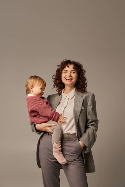 quality time, work life balance concept, happy woman in suit holding daughter and standing with hand in pocket on grey background, career and family, loving motherhood, confident leader  - Photo, Image
