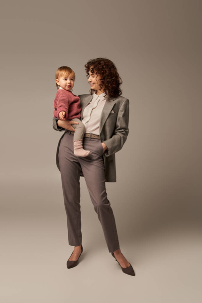 tiempo de calidad, concepto de equilibrio de la vida laboral, mujer sonriente en traje sosteniendo hija y de pie con la mano en el bolsillo sobre fondo gris, carrera y familia, maternidad amorosa, longitud completa  - Foto, imagen