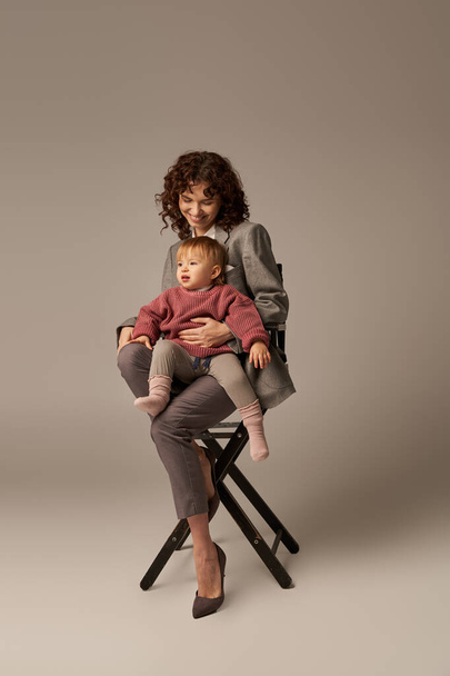 vyvážený životní styl, kariéra a rodina, kudrnatá žena v obleku sedí na židli s dcerou na šedém pozadí, profesionální úspěch, mateřství, multitasking, kvalitní čas, koncepce rovnováhy pracovního a soukromého života  - Fotografie, Obrázek