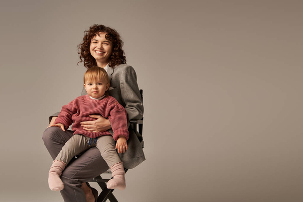 balanced lifestyle, career and family, curly businesswoman sitting on chair with child on grey background, professional success, motherhood, multitasking, quality time, work life balance concept  - Photo, Image