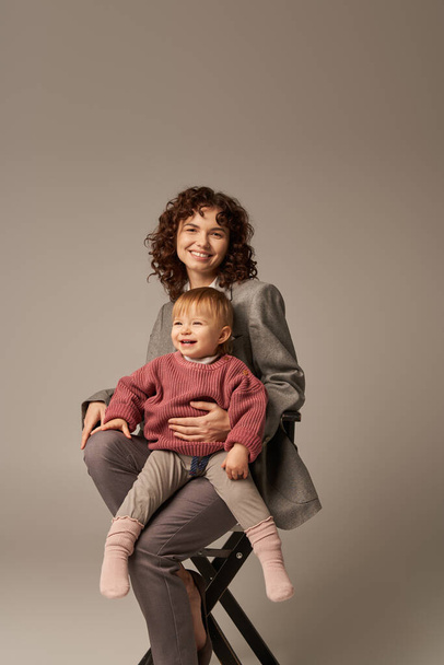balanced lifestyle, growth and family, happy businesswoman sitting on chair with happy child on grey background, professional success, motherhood, quality time, work life balance concept  - Foto, afbeelding