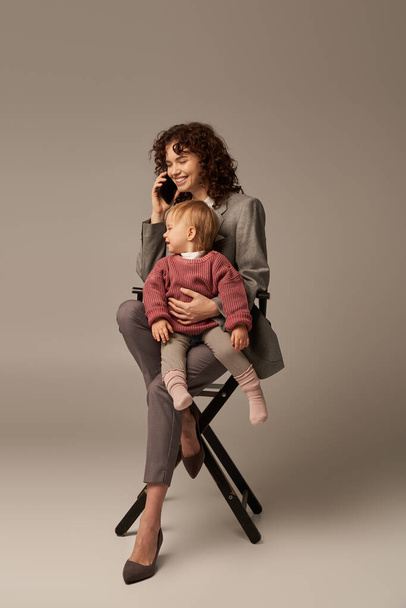 evenwichtige levensstijl, telefoongesprek, gelukkige zakenvrouw praten op smartphone en zitten op stoel met kind op grijze achtergrond, moederschap, multitasking, quality time, work life balance concept  - Foto, afbeelding