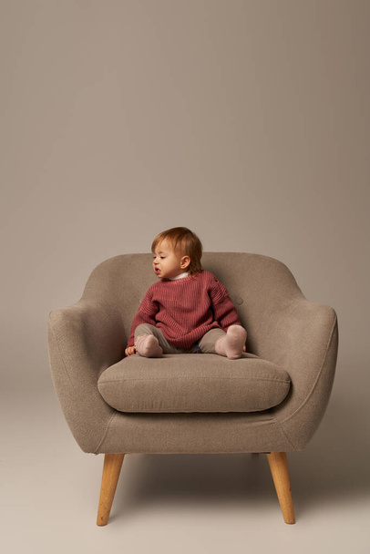 cute baby girl, toddler in casual attire sitting on comfortable armchair on grey background in studio, emotion, confused, innocence, little child, toddler fashion, stylish outfit, sweater  - Фото, зображення