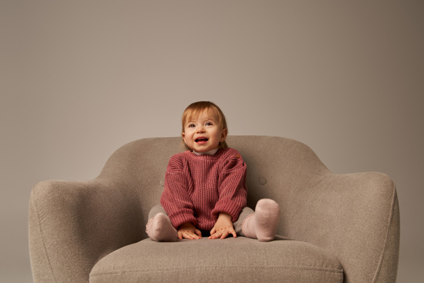 cute baby girl, toddler in casual attire sitting on comfortable armchair on grey background in studio, emotion, happiness, joy, innocence, little child, toddler fashion, stylish outfit, sweater  - Photo, Image