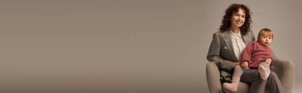 genitore lavoratore moderno, equilibrio tra lavoro e concetto di vita, donna riccia in giacca e cravatta seduta in poltrona con figlia bambino, sfondo grigio, madre e bambino allegri, multitasking, tempo di qualità, banner  - Foto, immagini