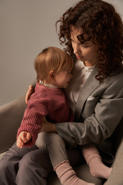 modern working parent, balancing work and life concept, curly woman in suit sitting in armchair with toddler daughter, calming crying baby, grey background, mother and child, multitasking  - Photo, Image