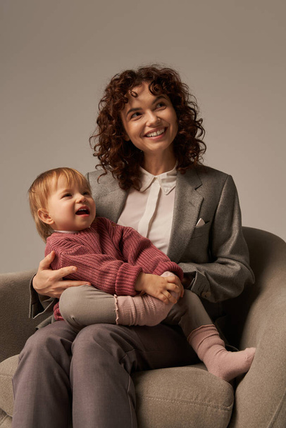 modern working mother, balancing work and life concept, happy businesswoman in suit sitting with toddler daughter on armchair, engaging with child, grey background, family relationships  - Foto, afbeelding