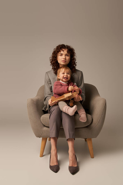 career and family, balancing work and life concept, businesswoman in suit sitting on armchair with toddler daughter, playing with wooden biplane, grey background, motherhood, full length  - Photo, Image