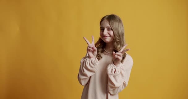 Blondýny teenager dívka 12 let v béžových šatech je šťastná a usmívá se a ukazuje vítězství znamení na žlutém pozadí ve studiu. dospívající dívka s úsměvem a ukázat dva prsty, vítězství gesto. - Záběry, video