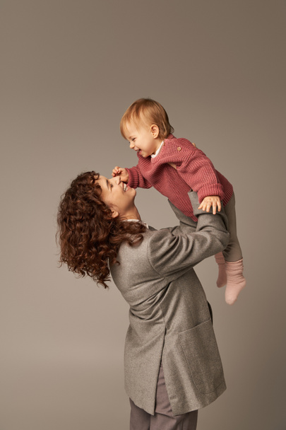 pracující matka, rodičovství a kariéra, veselá podnikatelka zvedající svou batolata dceru na šedém pozadí, koncept harmonie pracovního života, milující mateřství, kvalitní rodinný čas  - Fotografie, Obrázek