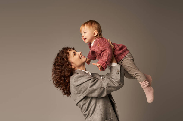 positivity, working mother, parenting and career, cheerful businesswoman lifting her happy toddler daughter on grey background, work life harmony concept, loving motherhood, quality family time  - Fotó, kép