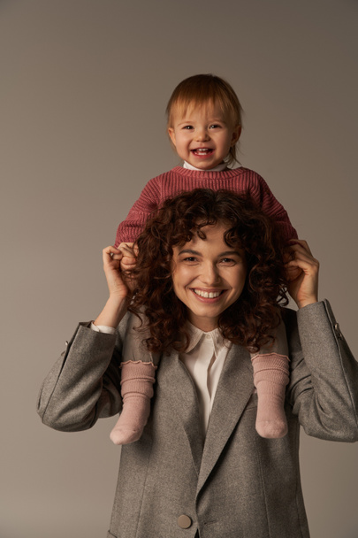 motherhood, work life balance, cheerful working mother with kid on shoulders, woman and happy child on grey background, work life harmony concept, loving motherhood, quality time  - Photo, Image