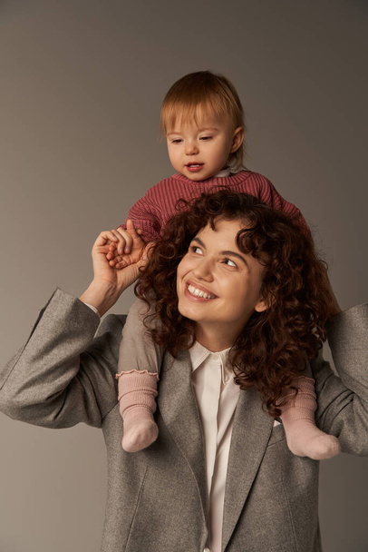 motherhood, kid on shoulders, work life balance, cheerful working mother, woman and happy daughter on grey background, work life harmony concept, loving motherhood, quality family time  - Foto, Imagem
