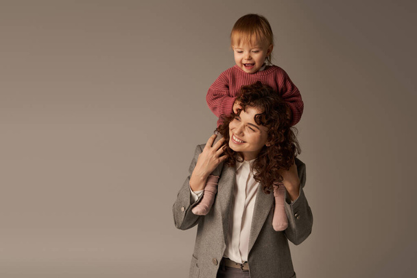 motherhood, kid on shoulders, parenting and career, curly businesswoman and happy daughter on grey background, work life harmony concept, loving motherhood, quality family time, work life harmony - Fotografie, Obrázek