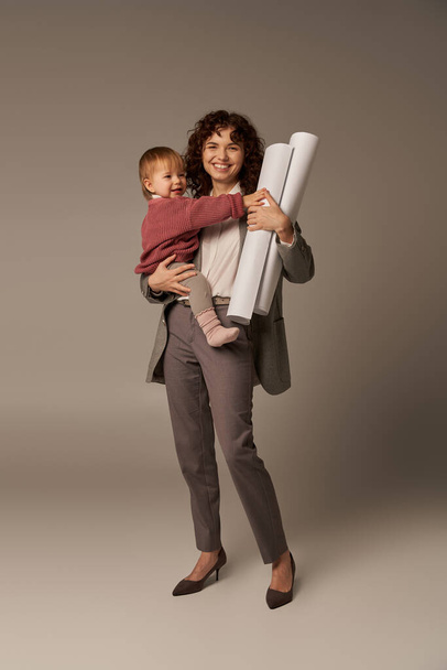 многозадачная женщина, профессиональные достижения, управление временем, счастливая мать, держащая в руках дочь и бумажные чертежи на сером фоне, построение успешной карьеры, полная длина  - Фото, изображение