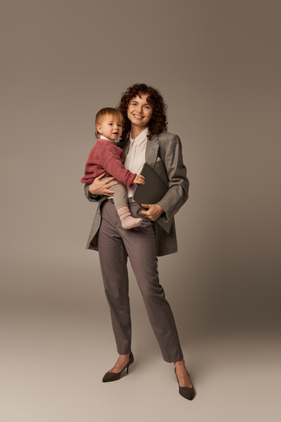 professional aspirations, modern parenting, work life harmony, happy working mother holding laptop and daughter on grey background, time management, self-confidence, full length  - Foto, immagini