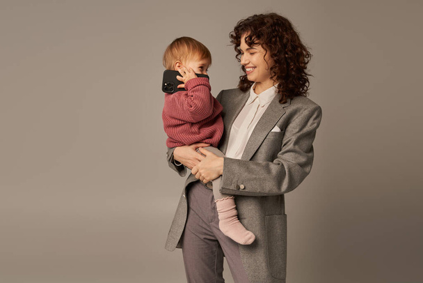 nowoczesne rodzicielstwo, budowanie udanej kariery, godzenie pracy z życiem, radosna matka trzymająca w ramionach córeczkę malucha ze smartfonem na szarym tle, macierzyństwo  - Zdjęcie, obraz