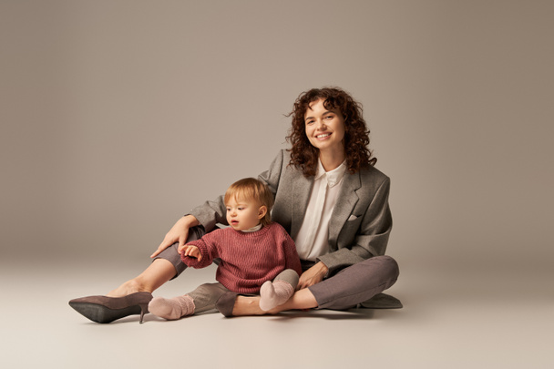 career and family, balancing between work and life, cheerful mother with curly hair hugging toddler daughter on grey background, quality time, modern parenting, businesswoman, full length   - Photo, Image