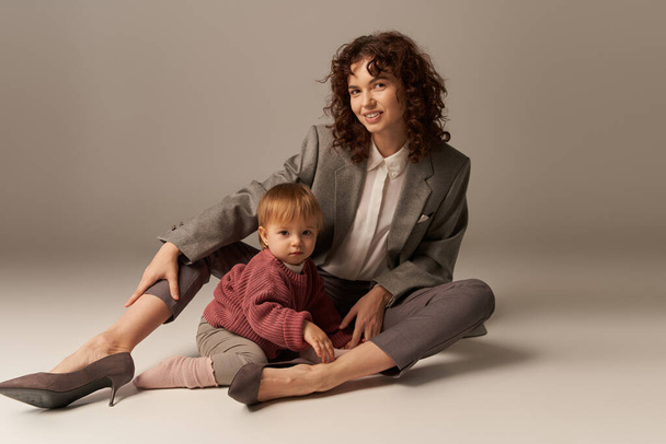 parenting and family, balanced lifestyle, cheerful mother with curly hair hugging toddler daughter on grey background, quality time, modern woman, businesswoman, loving motherhood   - Photo, Image