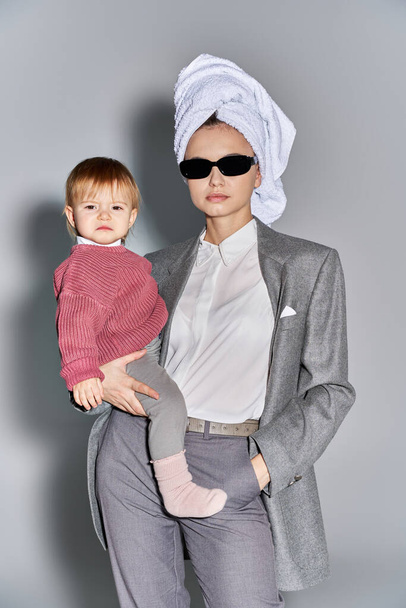 Multitasking, Frau mit Sonnenbrille, Kleinkind im Arm und mit Handtuch auf dem Kopf, Balanceakt zwischen Arbeit und Leben, ermächtigte Frau in formeller Kleidung auf grauem Hintergrund  - Foto, Bild