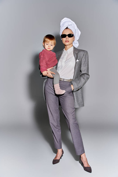 multitasking, woman in sunglasses holding in arms toddler daughter and standing with towel on head, balancing between work and life, empowered woman in formal wear on grey background, full length  - Photo, Image