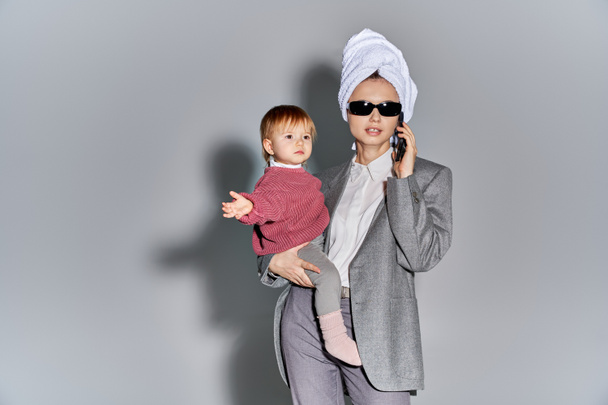 multitarea, mujer en gafas de sol sosteniendo en brazos hija niño y de pie con toalla en la cabeza, equilibrando el estilo de vida, mujer de negocios en ropa formal hablando en el teléfono inteligente sobre fondo gris  - Foto, imagen
